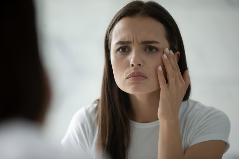 Close up head shot unhappy young woman wearing white t-shirt checking skin, looking in mirror, standing in bathroom, worried about mimic wrinkle or acne, touching face, skincare and treatment; Shutterstock ID 2173366879; purchase_order: ajanet; job: ; client: ; other: